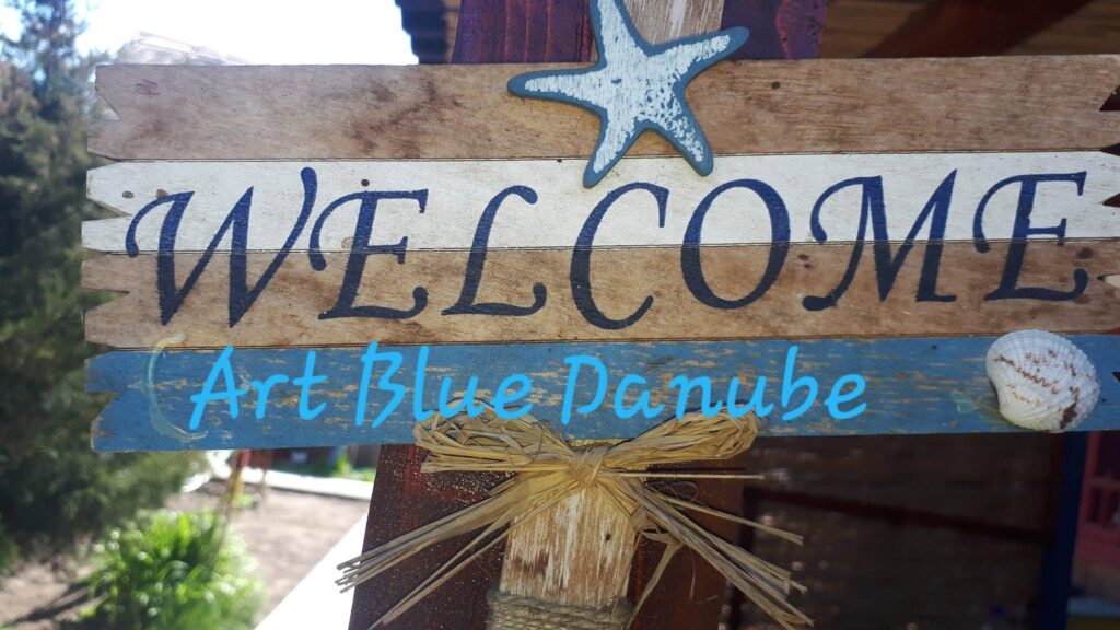 Firmă din scânduri văpsite pe care scrie Welcome și Art Blue Danube, decorată cu o stea de mare și o scoică, la intrarea în curte