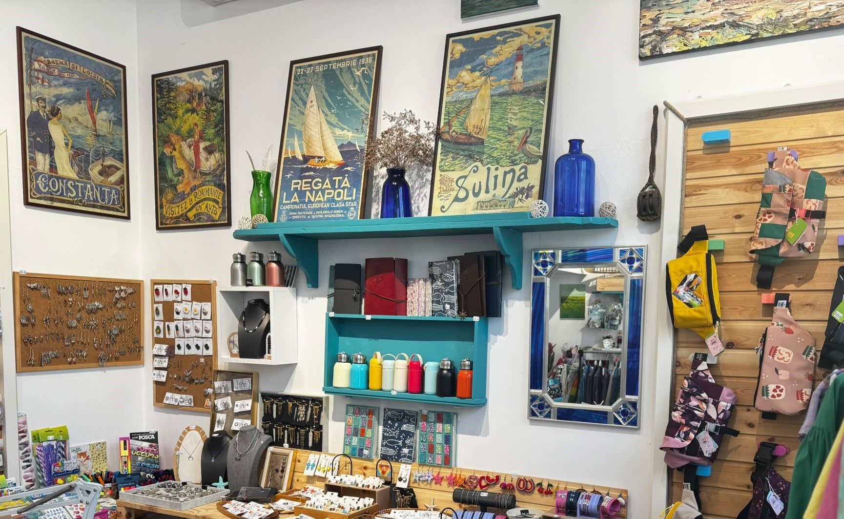 Interior de atelier-boutique cu afișe vechi de turism înrămate, rafturi turcoaz cu sticle albastre și standuri cu bijuterii și obiecte făcute de mână