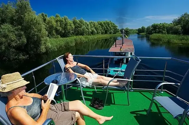 Două femei odihnindu-se pe punte, una citind, pe hotelul plutitor care înaintează pe un canal mărginit de sălcii în Delta Dunării