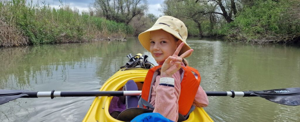 Copil cu pălărie și vestă de salvare portocalie, într-un caiac galben pe un canal mărginit de stuf, făcând semnul victoriei