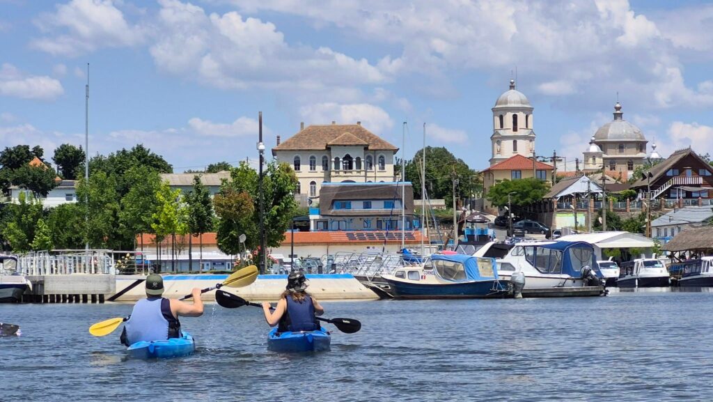 Doi caiaciști vâslind pe lacul Razim, cu portul, bărcile și turlele bisericii din Jurilovca în fundal
