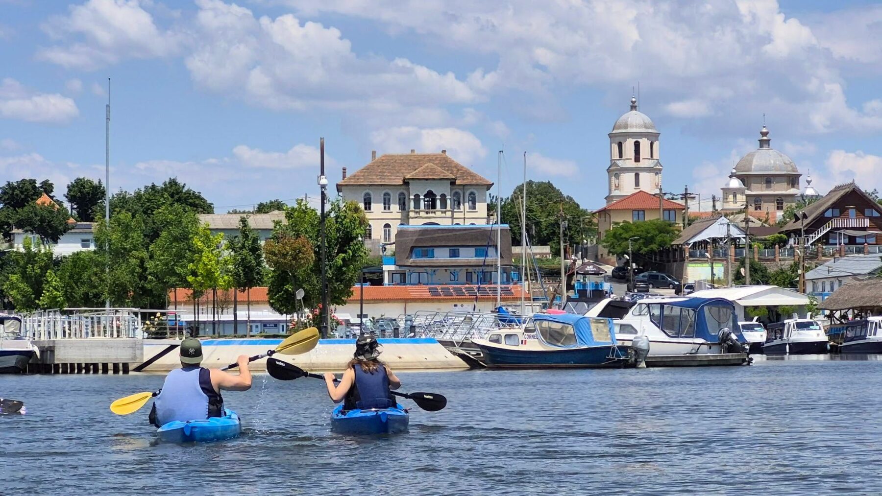 Doi caiaciști vâslind pe lacul Razim, cu portul, bărcile și turlele bisericii din Jurilovca în fundal
