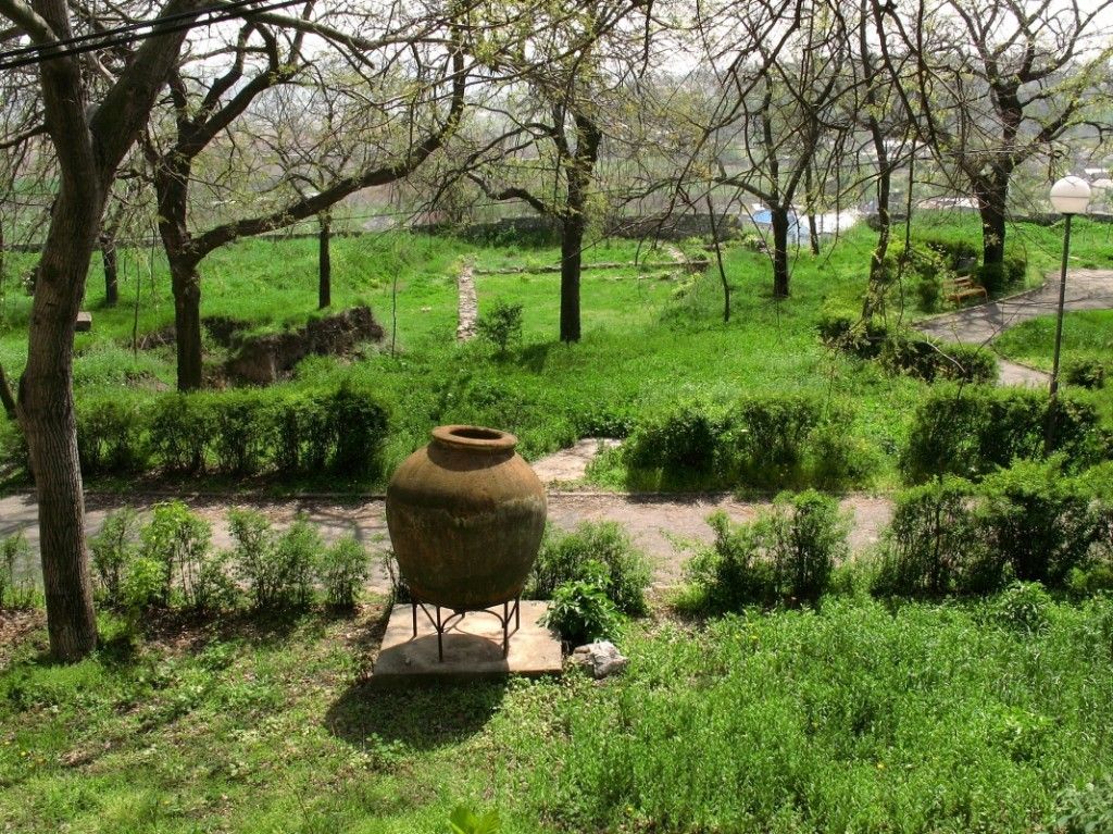 Amforă mare de lut expusă pe un suport de fier în parcul arheologic de pe Dealul Monumentului din Tulcea, cu urmele zidurilor cetății Aegyssus și cu pomi înverziți în spate