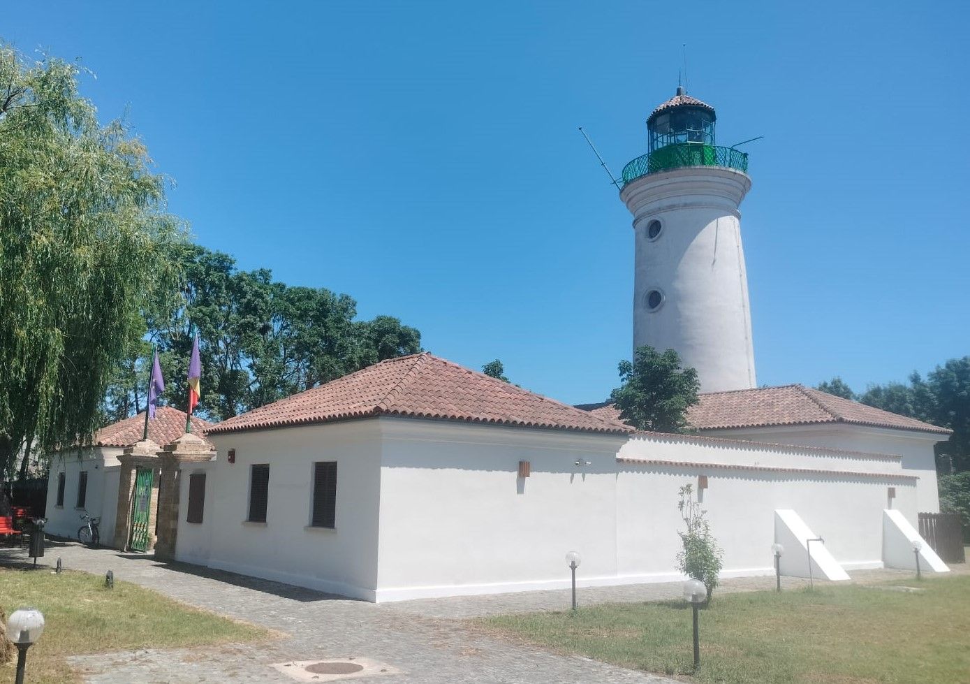 Turnul alb al Farului Vechi din Sulina, cu felinarul verde în vârf, ridicându-se deasupra clădirilor văruite și a acoperișurilor de țiglă roșie din jurul curții interioare