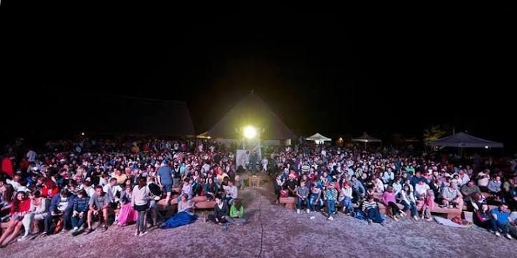 Mulțime numeroasă așezată pe nisip, noaptea, în fața ecranului cinematografului în aer liber de la Sfântu Gheorghe, cu corturile festivalului luminațe în spate