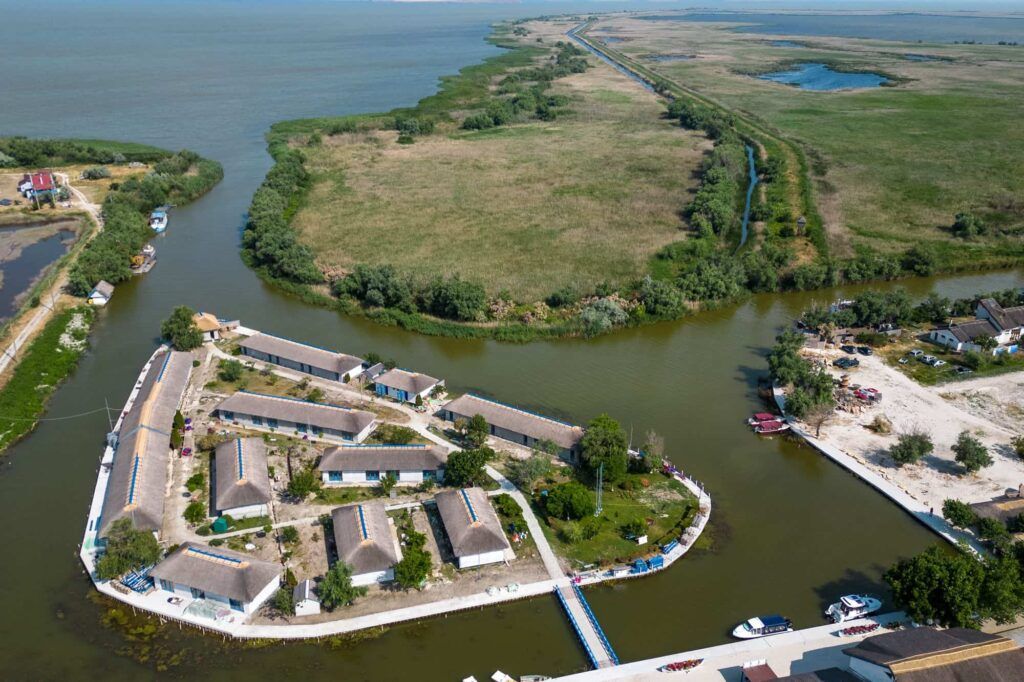 Vedere de sus asupra căsuțelor cu acoperiș de stuf de la Gura Portiței, așezate pe fâșia de pământ dintre lagună și Marea Neagră, cu pontonul și bărcile alături