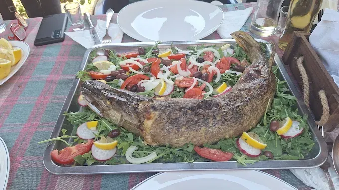 Pește mare copt întreg, servit pe tavă cu roșii, ceapă, măsline, lămâie și rucola, pe o masă așezată pentru mai multe persoane