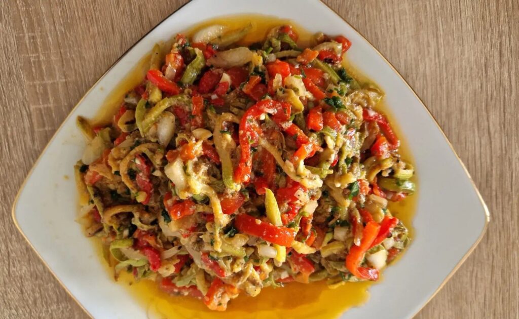 Farfurie albă cu salată de ardei copți tăiați fâșii, cu roșii, ceapă și ulei, văzută de sus pe o masă de lemn