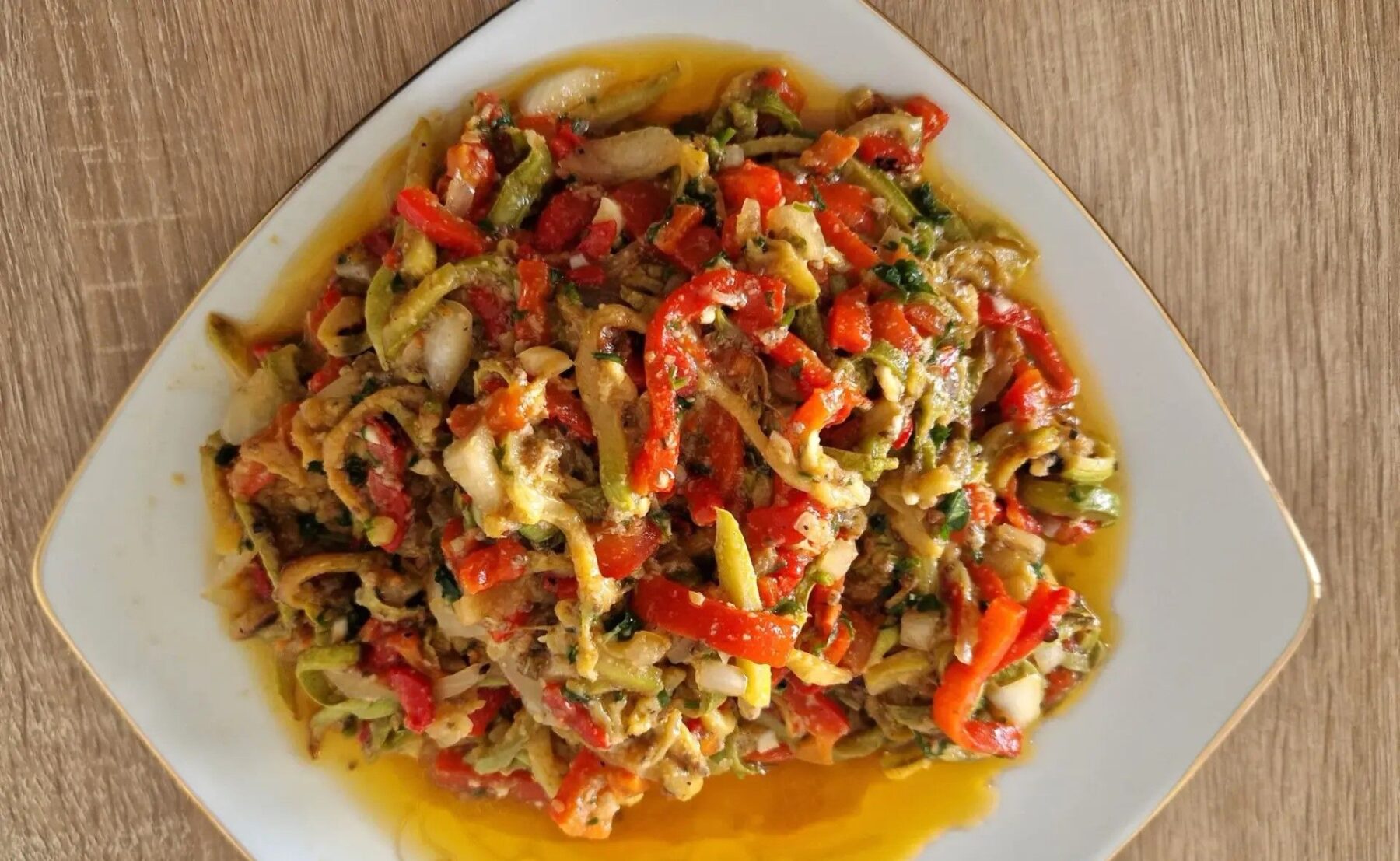 Farfurie albă cu salată de ardei copți tăiați fâșii, cu roșii, ceapă și ulei, văzută de sus pe o masă de lemn