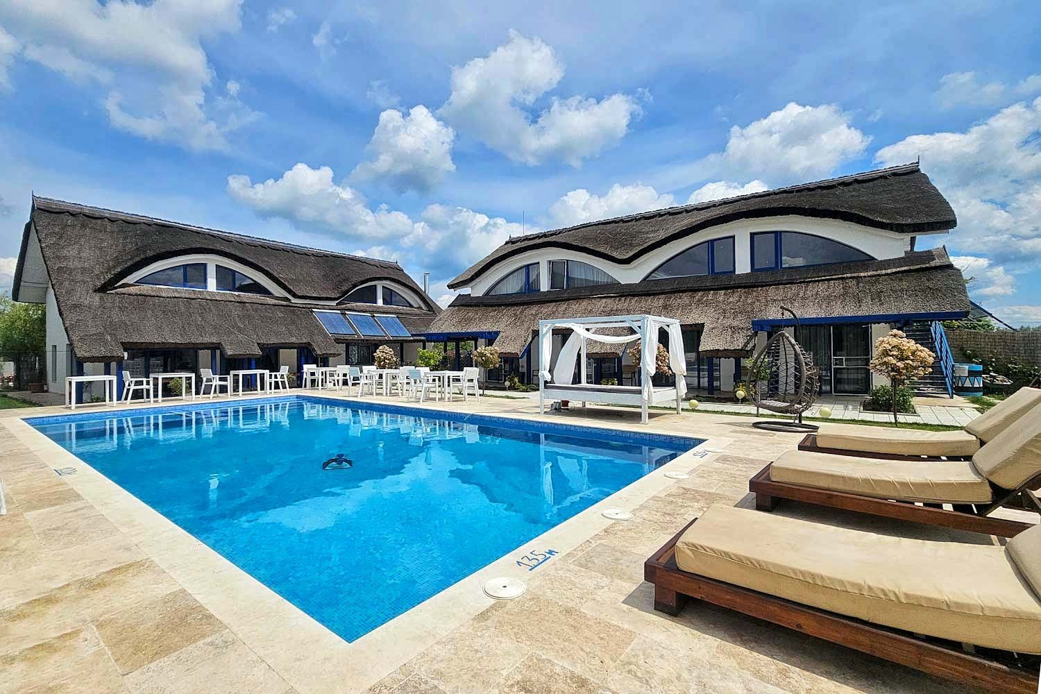 Două clădiri de pensiune cu acoperișuri ondulate din stuf și șezlonguri în jurul unei piscine, la Murighiol