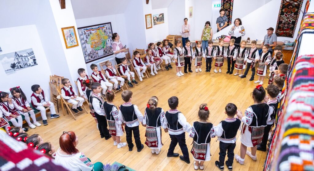 Grup de copii în costume populare așezați și stând în cerc într-o sală a muzeului, cu țesături și covoare tradiționale pe pereți