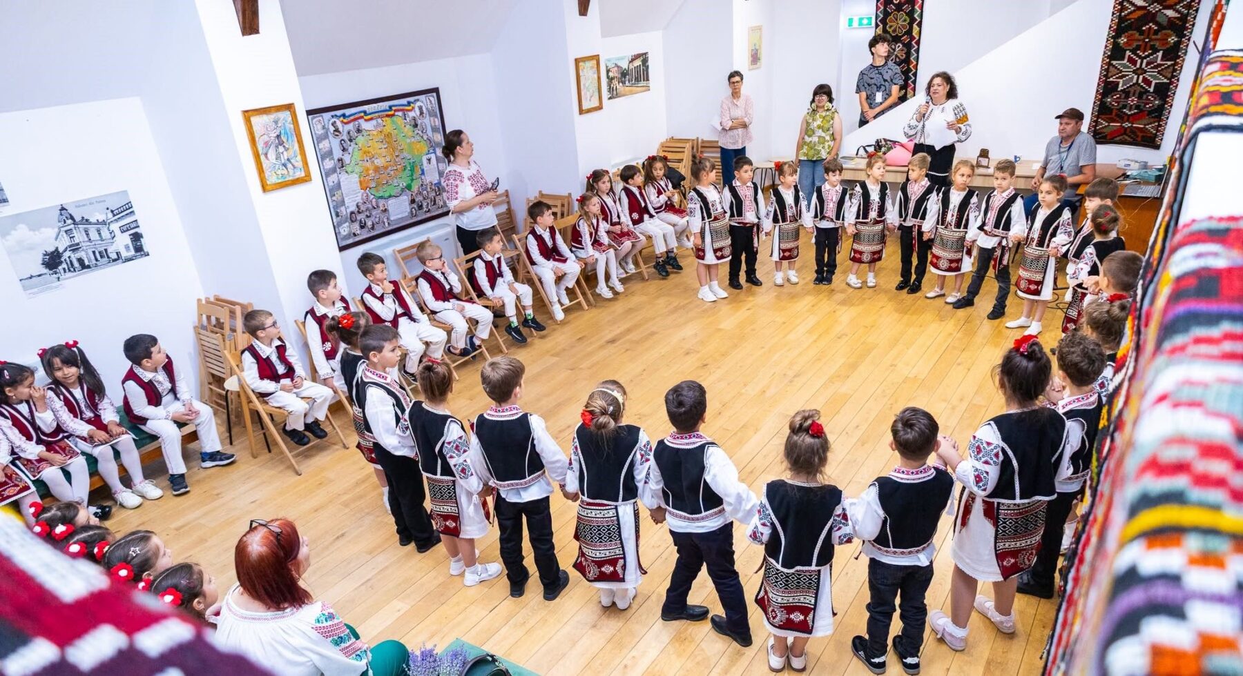 Grup de copii în costume populare așezați și stând în cerc într-o sală a muzeului, cu țesături și covoare tradiționale pe pereți