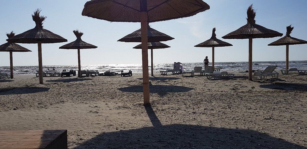 Umbrele de stuf și șezlonguri pe nisipul plajei de la Sulina, cu marea în contralumină și cățiva oameni la mal