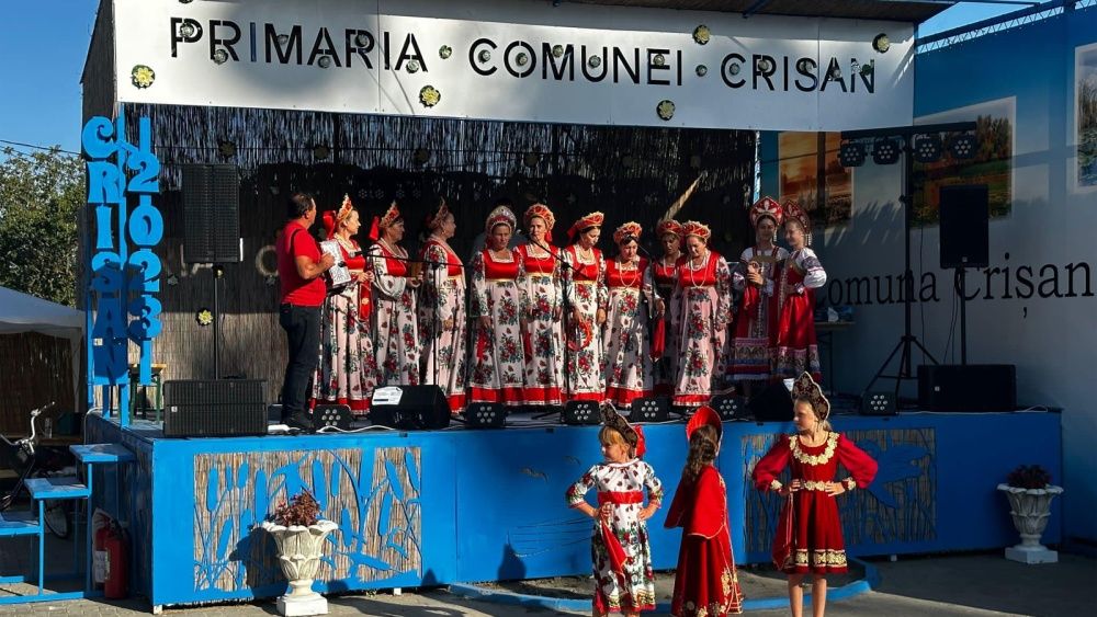 Cor de femei în costume lipovenești roșu cu alb pe scena Primăriei comunei Crișan, cu trei fetițe în costum popular în fața scenei