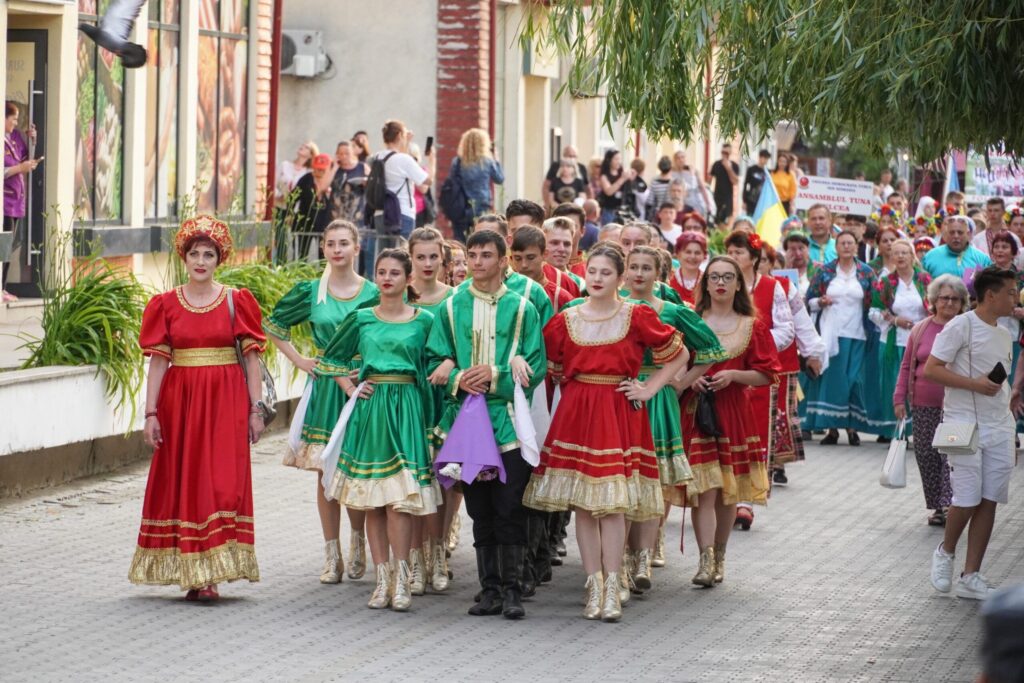 Alai de tâneri în costume populare roșii și verzi defilând pe o stradă din Sulina, urmați de un grup lung de participanți și de spectatori