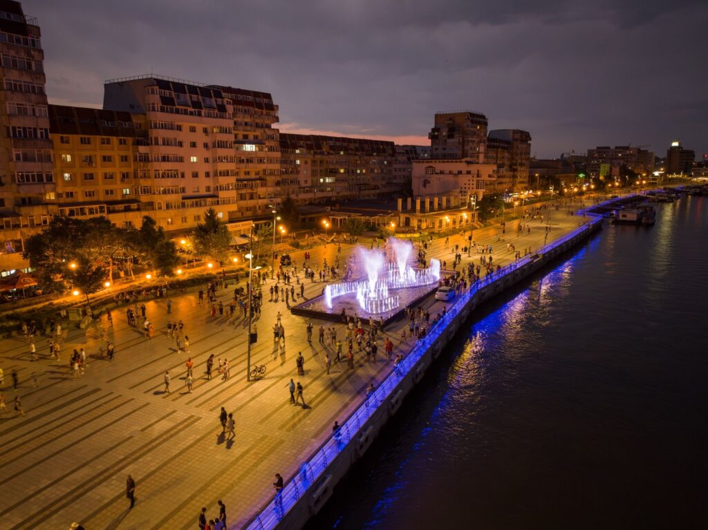 Faleza Tulcei seara, văzută de sus: fântâna arteziană luminată, promenada plină de oameni și malul Dunării cu lumini albastre